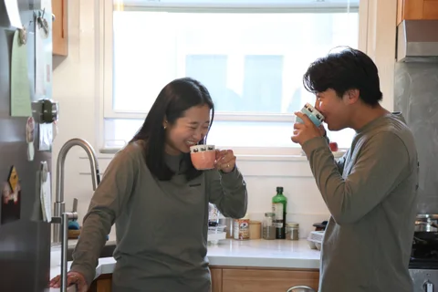 Nam-Hee and Hiroo drinking from cups in a kitchen.